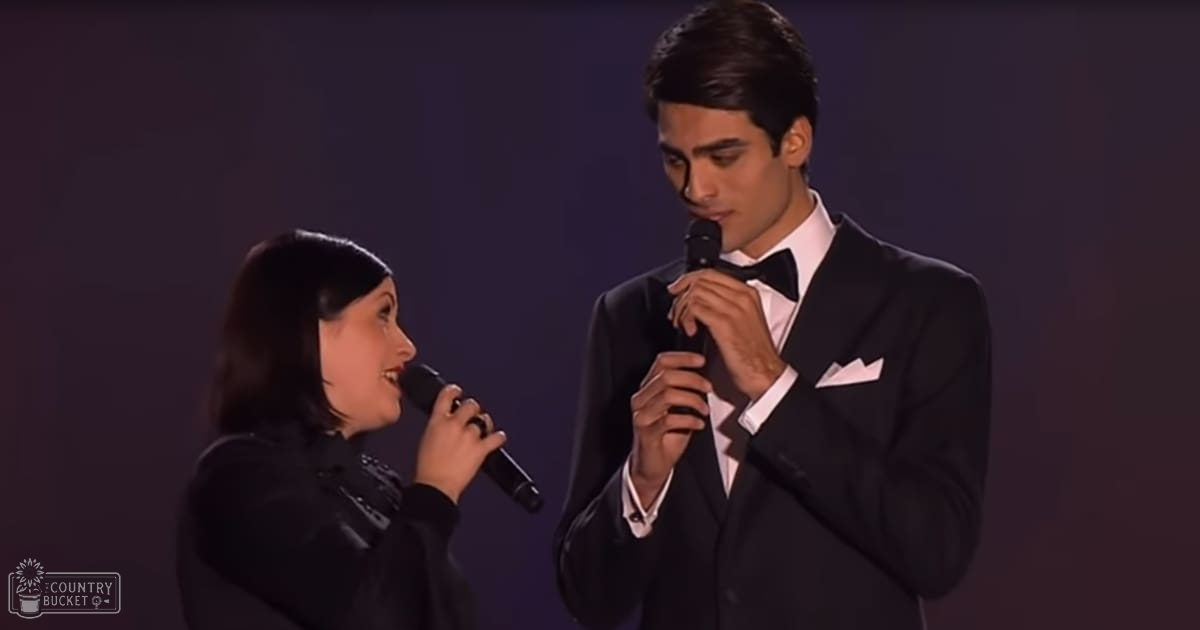 A duet performance scene with two young adult performers, one in a modern black top and the other in an early-to-mid 20th-century style tuxedo, set against dark curtains on stage under dramatic lighting.