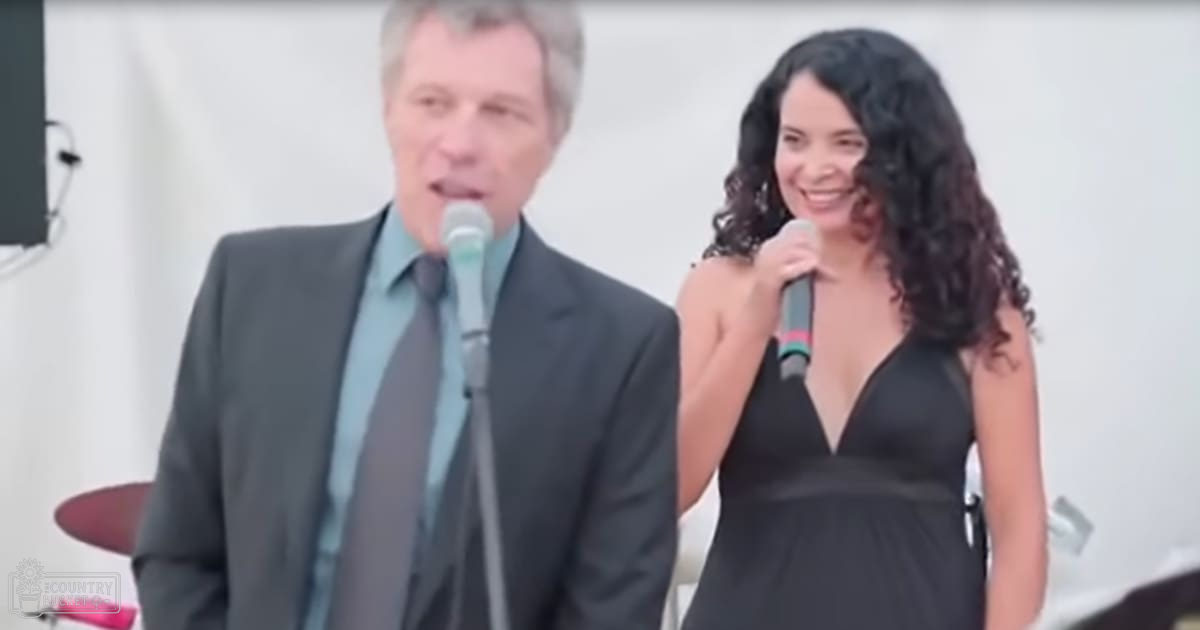 Middle-aged male singer in dark gray suit with light blue dress shirt and dark tie, accompanied by a young adult female in black sleeveless dress, microphone stand visible, drum set partial view, plain white background suggesting outdoor event setting.