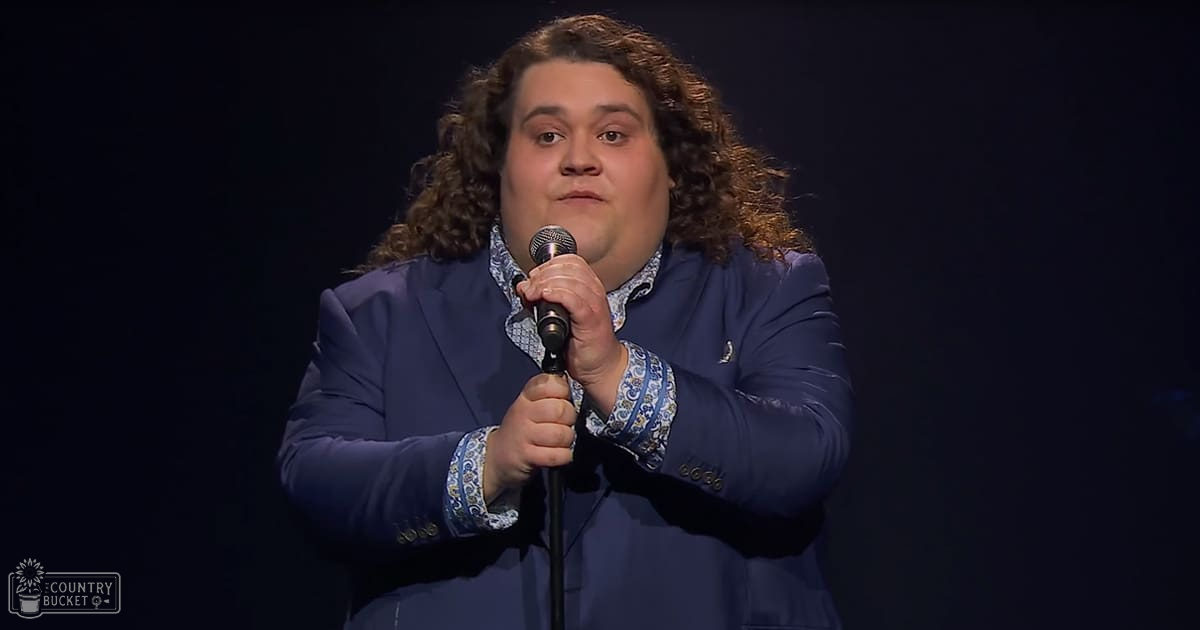 A middle-aged male comedian in a dark blue blazer performs stand-up comedy on stage under dim lighting at an indoor venue.