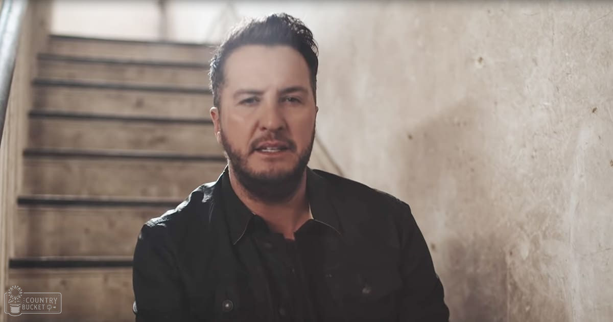 An adult male in a relaxed, modern casual button-up shirt with no visible pattern, set against an aged rustic wooden staircase and worn walls illuminated by natural light.