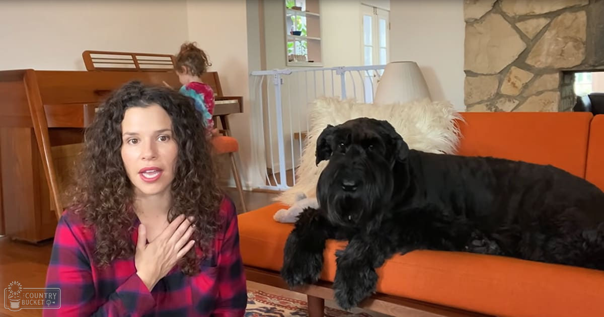 A young adult to middle-aged female in a living room with orange sofa, stone wall paneling, piano, and natural light suggesting daytime spring or summer.