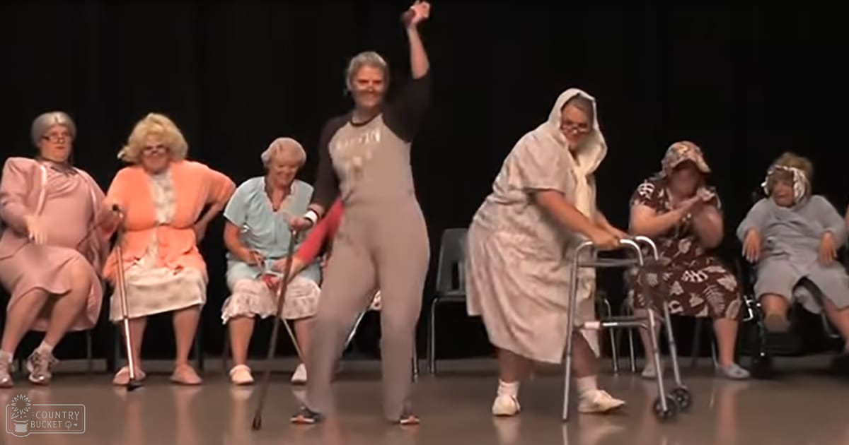 A middle-aged woman performing a dance routine with older adults seated behind her indoors under artificial lighting, wearing a light gray or beige form-fitting bodysuit.