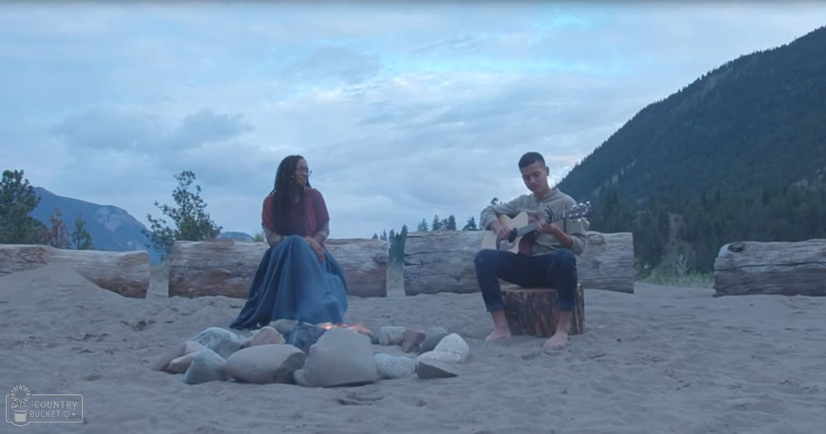 A young adult male and female enjoy a beachside campfire scene with scattered driftwood logs and trees in the background under an overcast sky.
