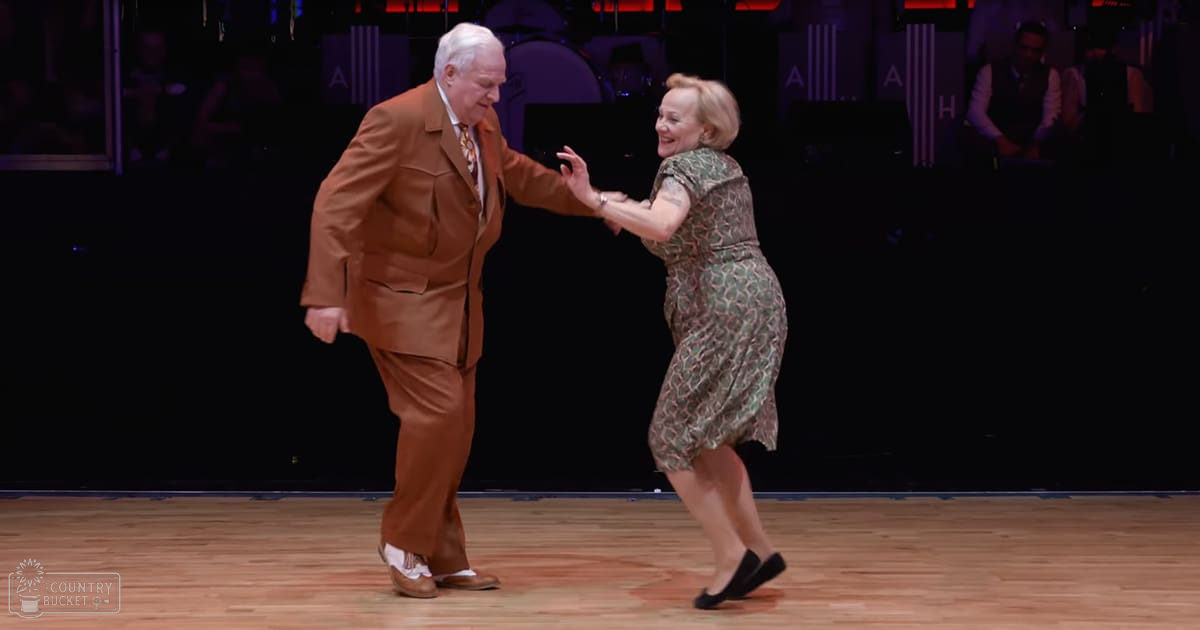 An older adult male and female couple dancing indoors, the man wearing a brown suit with white dress shoes, the woman in a green patterned dress with black slip-on shoes.