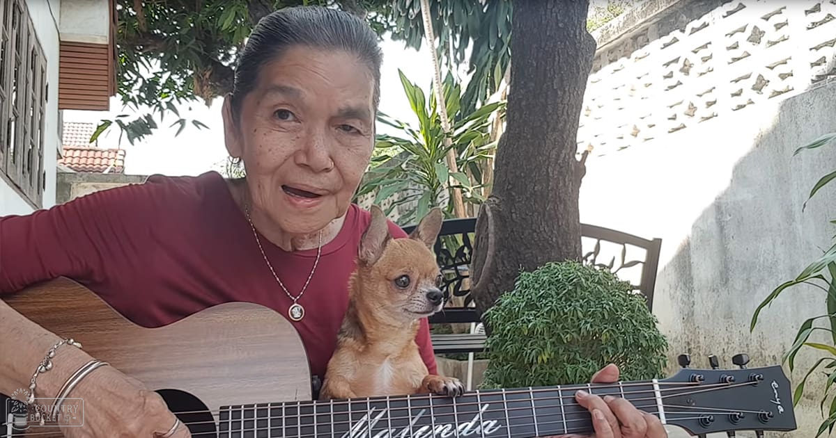 An older adult woman plays a guitar outdoors in a residential area with trees, plants, and a concrete wall. A Chihuahua dog is nearby.