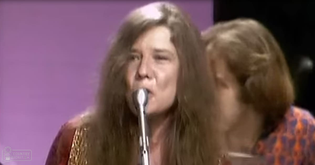 Adult female performer singing into a microphone, wearing a reddish-brown dress with gold accents, accompanied by another adult female in an orange-blue patterned blouse; stage setting with solid purple backdrop and artificial lighting.