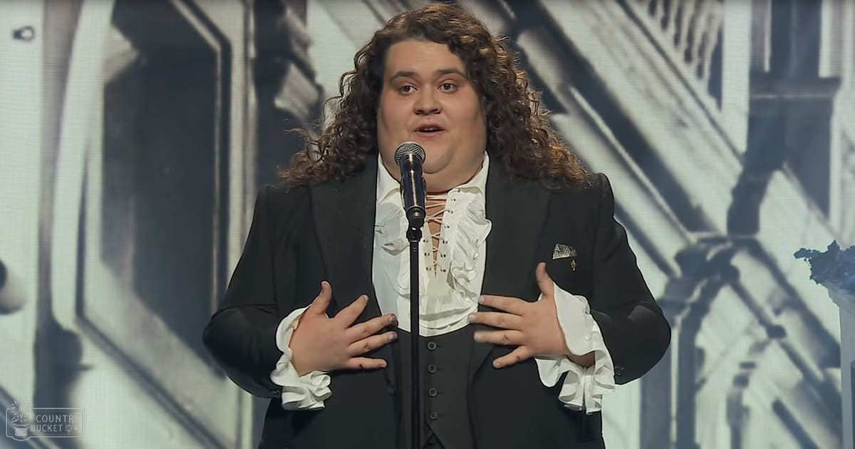 A young adult male in a black suit performs at an awards show, gesturing with hands near chest against backdrop of abstract designs and stage lighting.