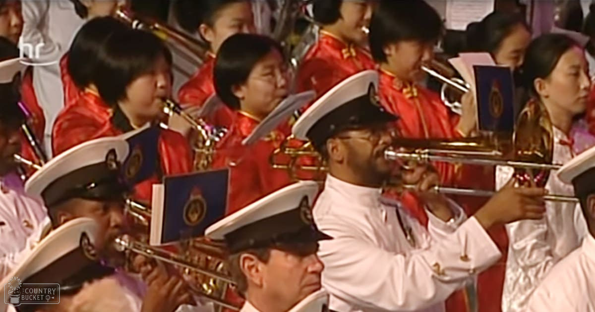 A military band performance in an indoor setting with adult males and females wearing white uniforms and red jackets, playing trumpets and saxophones. The venue is possibly a concert hall or auditorium during the evening.