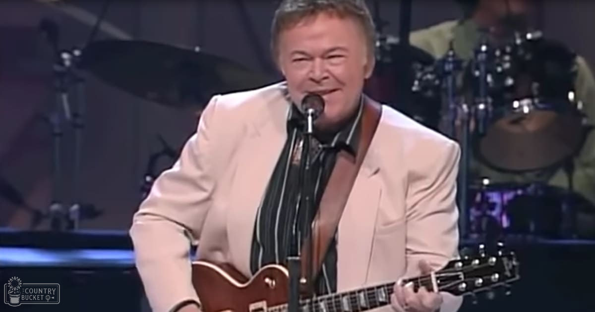 A middle-aged male performer in a white suit jacket and dark tie stands at a microphone stand, surrounded by musical instruments under stage lighting.