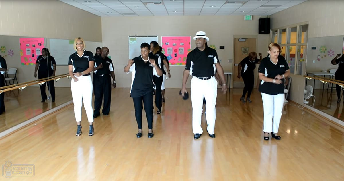 An indoor dance studio with a group of middle-aged and older adults engaged in a class, mirrors along walls, whiteboard visible, wooden floors, and posters on the wall.