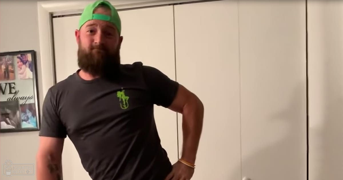An adult male in a fitted dark gray crew-neck T-shirt with short sleeves, wearing a green baseball cap backward and a gold bracelet on his left wrist, indoors near white wardrobe doors and framed pictures.