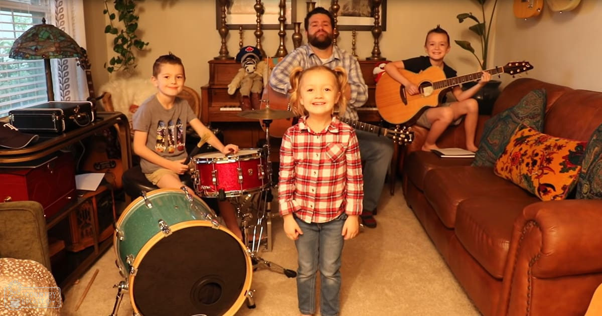A young girl in a red plaid shirt and blue jeans stands among other children, an adult male singer in a blue and white plaid shirt, and various musical instruments like drums and guitars. The setting is a cozy living room with patterned throw pillows, a drum set, and a soft natural light.