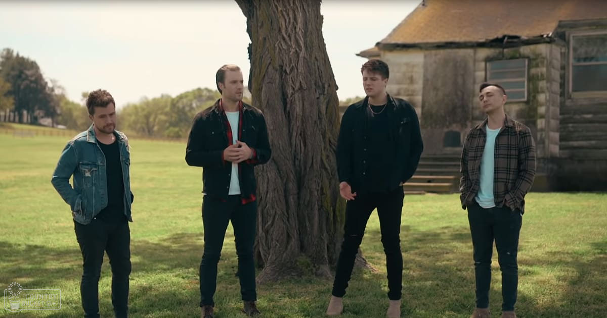 Four young adult males in a rural outdoor setting with grassy fields, trees, an old stone building, and clear sky suggesting late afternoon or early evening.