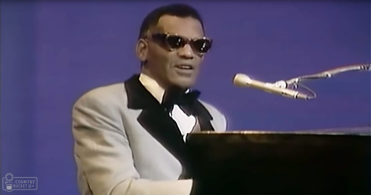 Adult male performer in a well-fitted light-colored suit jacket playing the piano on stage with a microphone nearby against a plain blue backdrop.