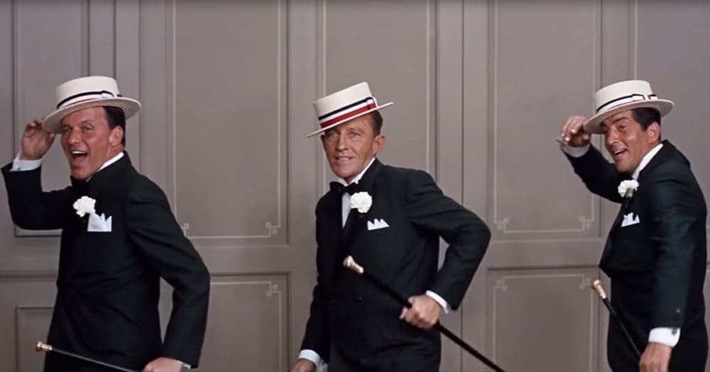 Adult males dressed in formal tuxedos with white boutonnieres, holding canes or hats, set against paneled walls suggesting a theater or dance hall interior.