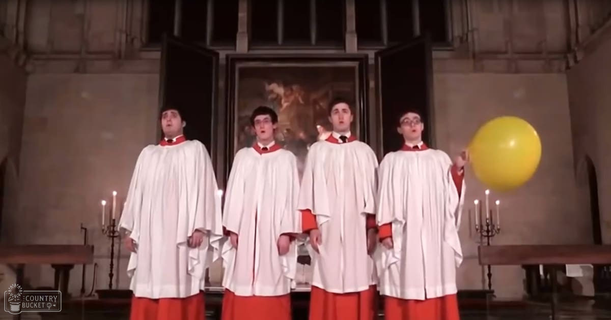 A group of young adult male singers in choir robes performing inside a church or cathedral with two lit candelabras, a yellow balloon, and religious artwork in the background.