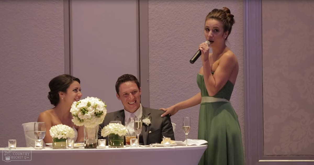 A young adult female in a strapless green wedding dress giving a speech at an indoor wedding reception, with the bride and groom seated at a long white table set for an event.