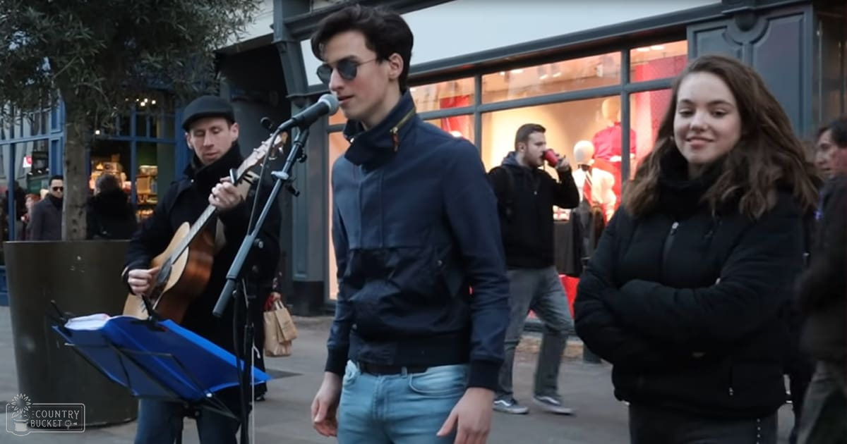 A young adult male singer wearing sunglasses and a dark blue zip-up jacket, performing on an urban street with sheet music and shopping bags visible.