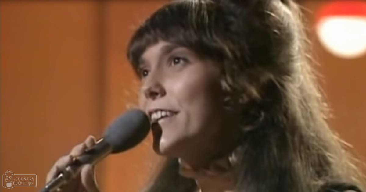 A young adult female sings into a microphone in an indoor setting with an orange wall and circular light fixture. The scene appears to be part of a performance or recording setup, with no clear focus on any specific object.