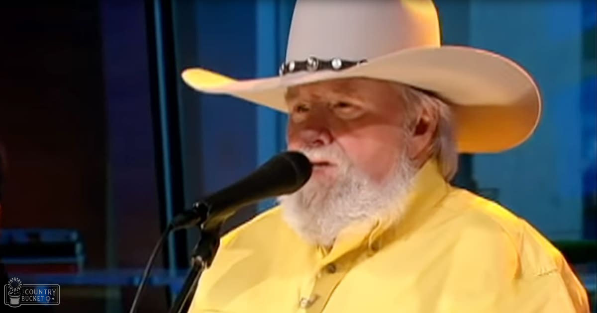 A middle-aged man wearing a bright yellow button-up shirt, wide-brimmed white cowboy hat with dark band, singing into a microphone on what appears to be an indoor stage or studio setup.