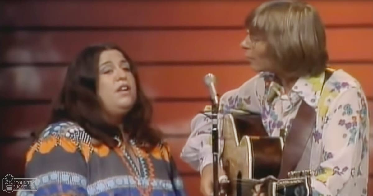 A musical performance with two adults on stage, one wearing a patterned blouse and beaded necklace, another in a floral shirt holding an acoustic guitar, set against red paneling suggestive of a theater or concert hall.