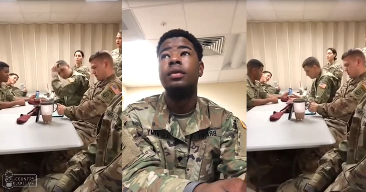 A young adult male in a camouflage-patterned military uniform sits at a white table with other individuals also in similar uniforms, surrounded by beige walls and fluorescent lighting.