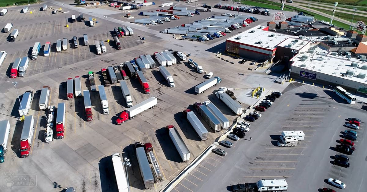 An outdoor truck stop parking lot with numerous parked trucks, cars, and RVs under clear daylight conditions.