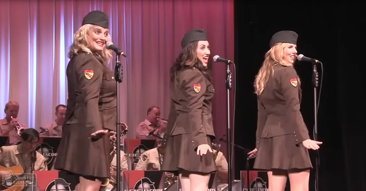 Three women in military-style uniforms perform on stage with an orchestra backdrop under evening lighting.