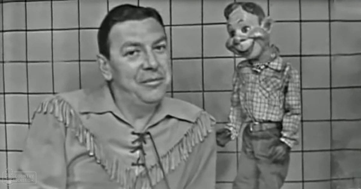 A middle-aged male in a Western-themed costume stands against a simple grid backdrop indoors under bright even lighting.