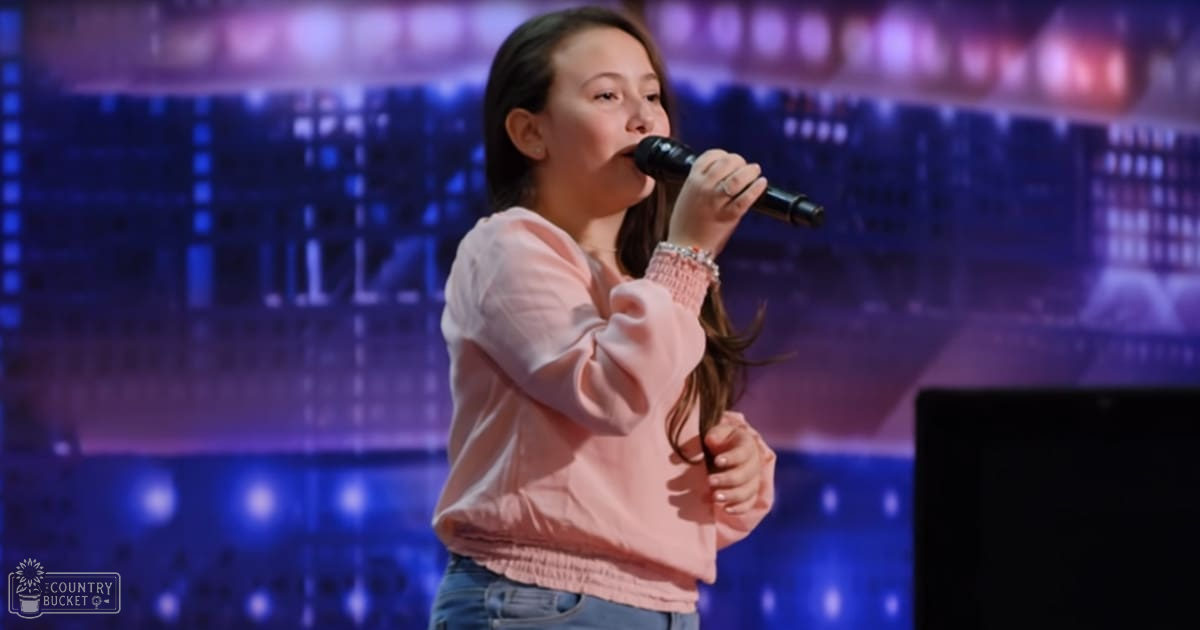 A young child wearing a light pink, long-sleeved top in a talent show setting, singing into a microphone.