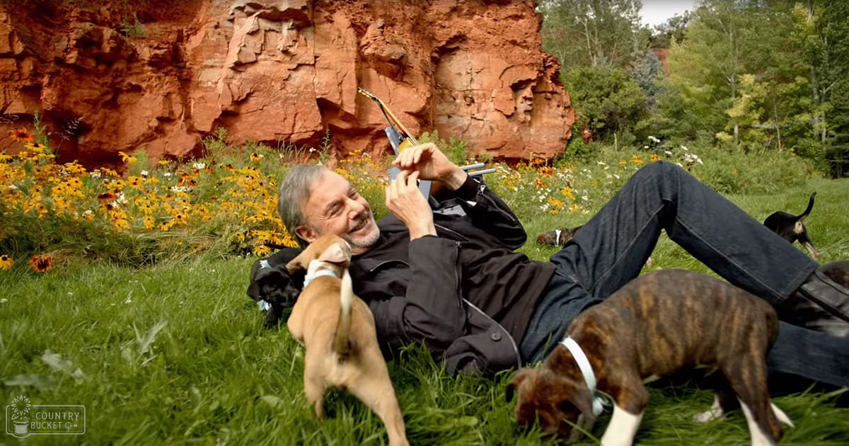 A middle-aged adult male wearing a black jacket outdoors near red rock formations and wildflowers, interacting with two light brown puppies.
