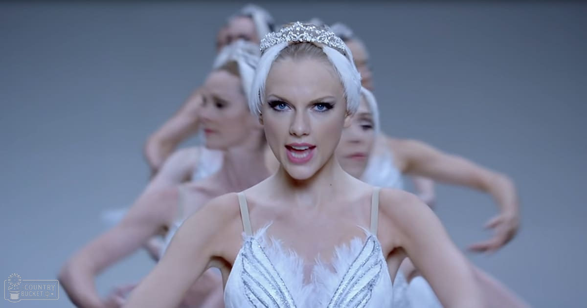 A young adult female wearing a tiara and white feathered dress stands in an indoor setting with multiple blurred individuals in similar outfits in the background, all under even artificial lighting.