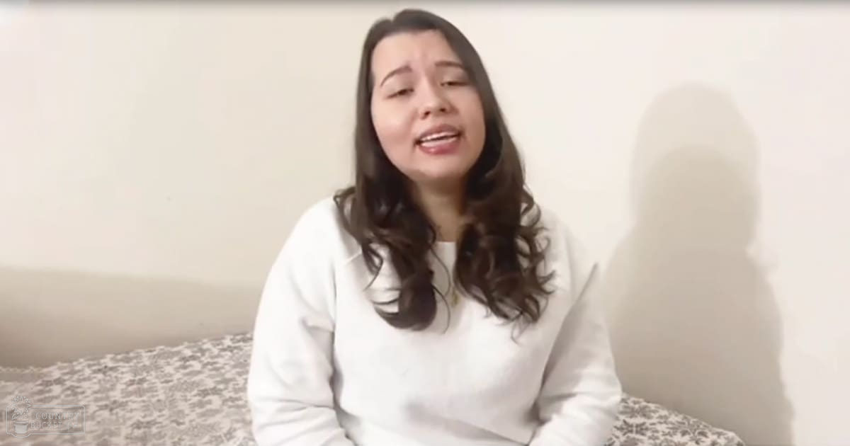 A young adult female in a relaxed indoor setting, wearing a white long-sleeved sweatshirt with neutral lighting and patterned bedding.