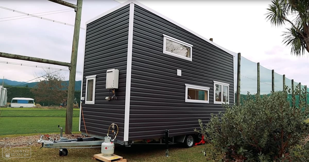 A rural scene with a tiny house, propane tank, white van, fence, green netting, wooden posts, bushes, grass, electrical poles, and window air conditioning unit; possibly spring or summer daytime with overcast skies.