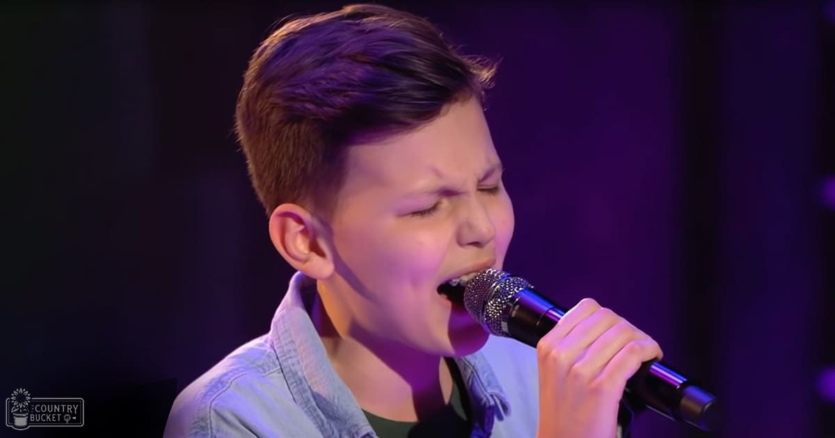 A young male child sings into a microphone, wearing a relaxed fit, light blue denim shirt with long sleeves in an indoor performance setting.