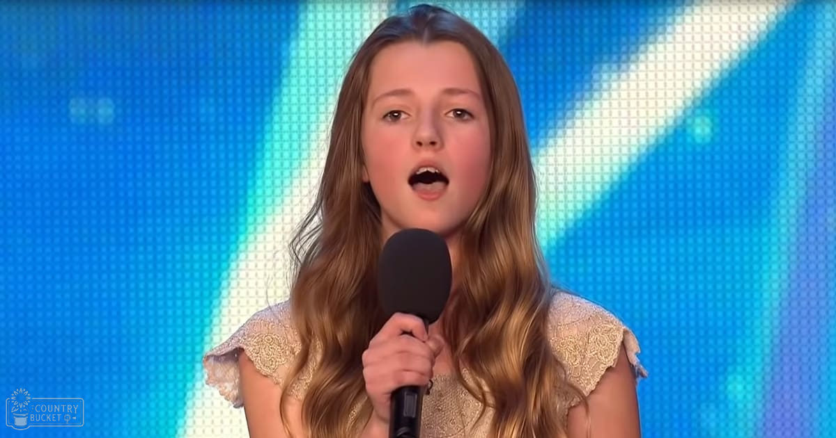 A young female child performing on stage, wearing a light-colored dress with ruffled sleeves and lace-like shoulders, holding a microphone under blue stage lighting.