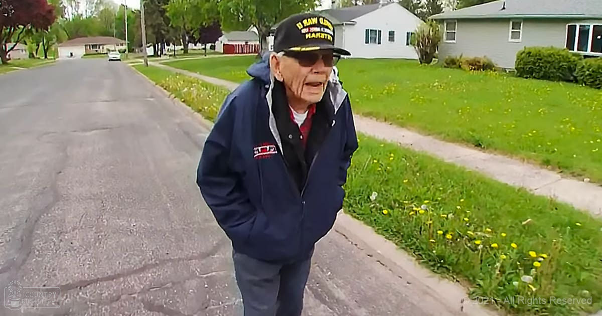 An older adult male wearing a dark blue jacket with '10 RAW CLAW' cap and sunglasses walks down a suburban residential street lined with houses, lawns, and sidewalks.