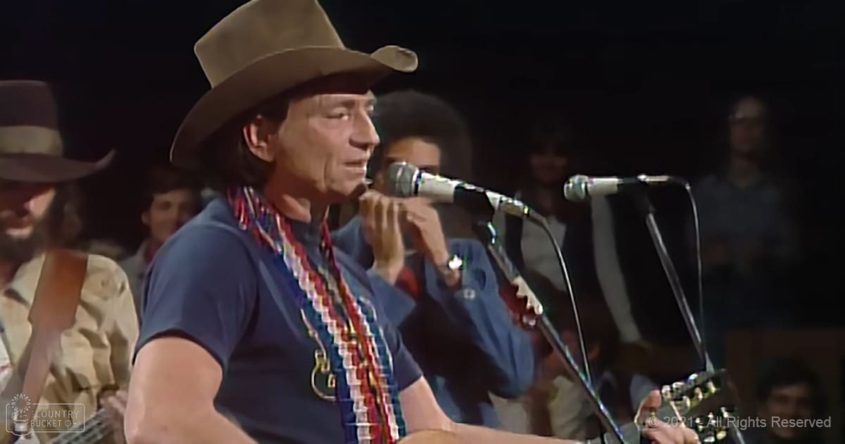 A middle-aged male singer wearing a dark blue t-shirt with a red and white patterned necklace, performing into a microphone at what appears to be a concert in an indoor venue with artificial lighting.