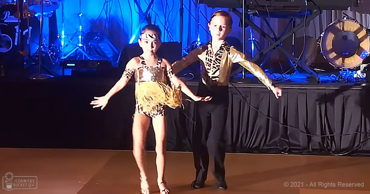 A dance performance scene at an indoor venue with children in leopard print leotards and a male performer in a black suit, dim blue stage lighting creating ambiance suitable for a performance.