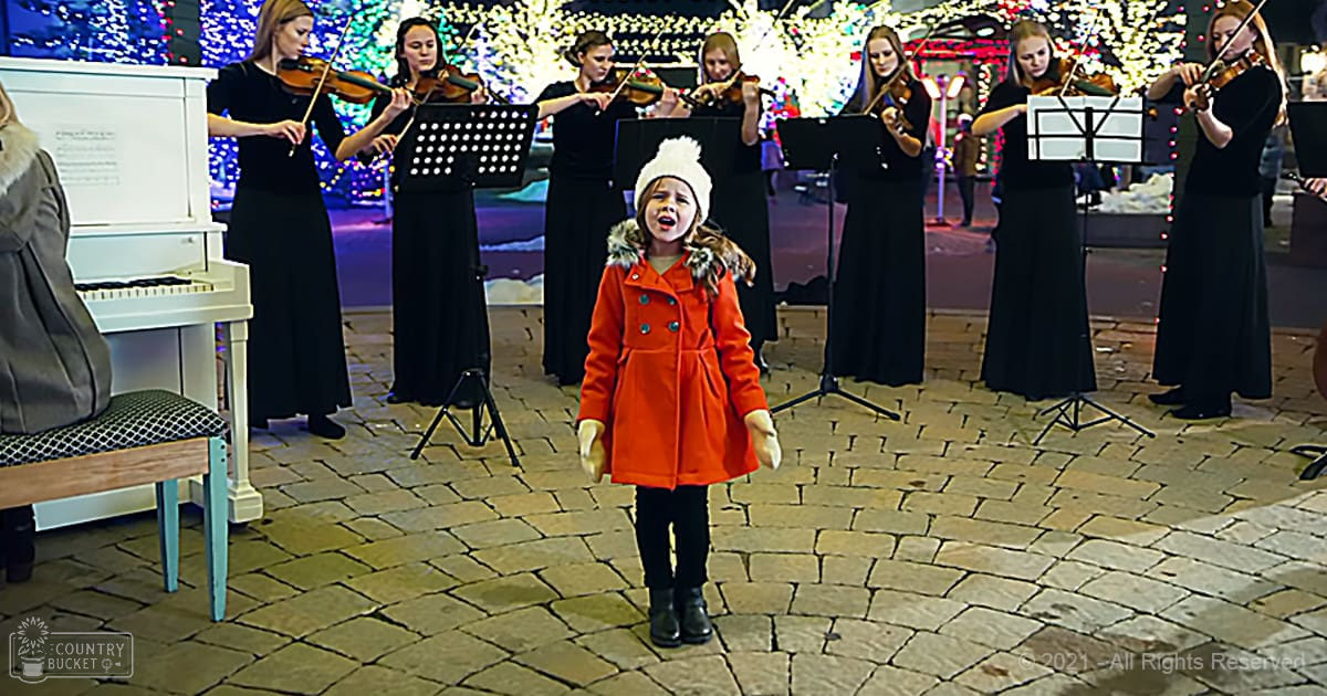 A young child wearing a red coat with fur trim stands outdoors at night during what appears to be a holiday season, surrounded by musicians in long black dresses and white hats performing under festive lights.