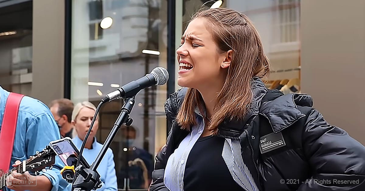 A young adult female singing outdoors in an urban setting with a male adult holding a guitar and another person in the background, wearing a blue jacket. A microphone stand and smartphone are visible.