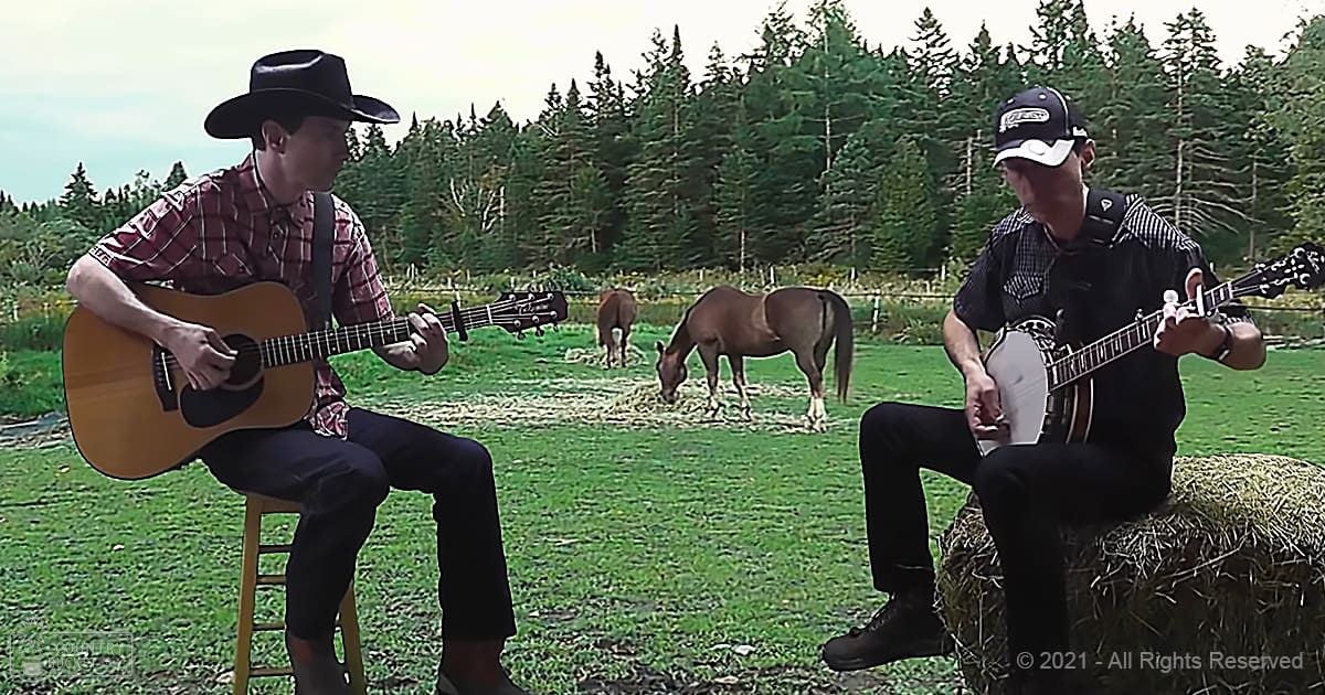 A musical duo in an outdoor rural setting with horses grazing, wearing casual attire and sitting on hay bales amidst green grass and trees.