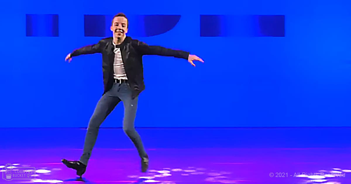 A young adult male dancing on a stage with a blue backdrop, wearing a black jacket over a white shirt and dark jeans.