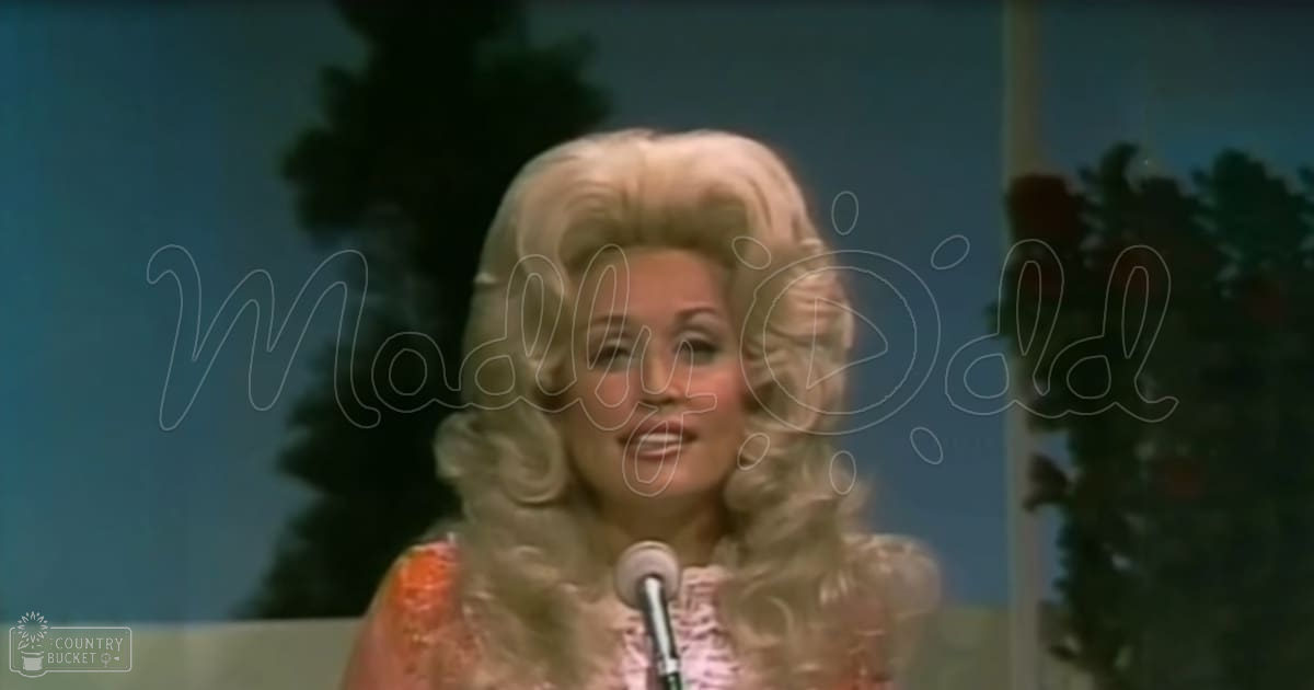 An adult female performer with a voluminous curly blonde wig, wearing an orange-toned patterned garment in front of dark green foliage suggesting an outdoor backdrop.