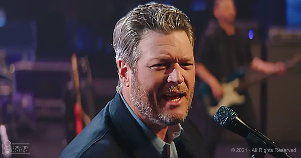 A middle-aged man, wearing a dark blue suit jacket and light gray shirt with a collar, performs on stage at an indoor music venue under dim spotlight lighting.