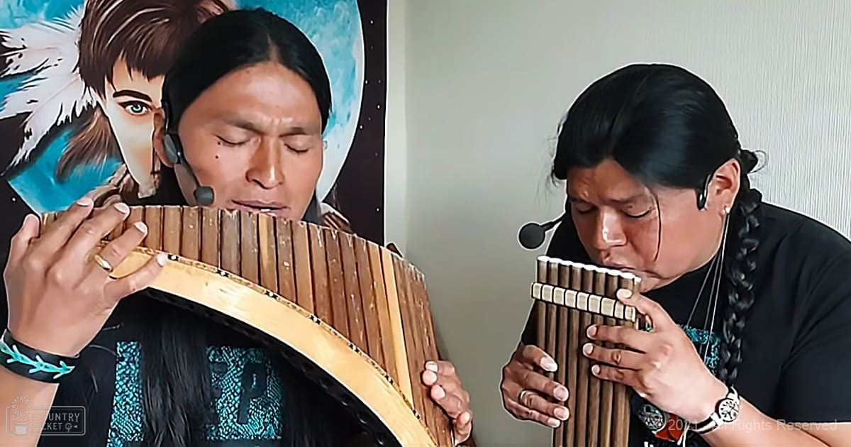 A musician playing a pan flute indoors, wearing black T-shirts with cultural patterns, surrounded by another individual holding similar instruments and a serene mural on the wall.
