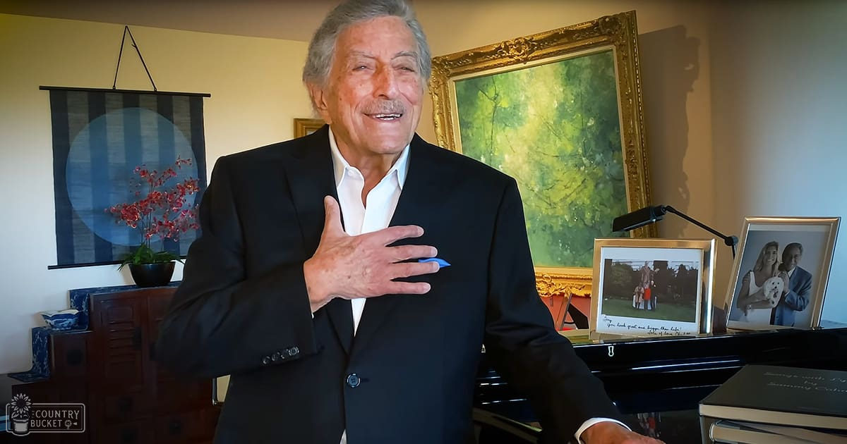 An older adult male wearing a black suit jacket with white shirt, standing indoors near a piano and framed photographs. The setting suggests a home office or living room with diffused natural light.