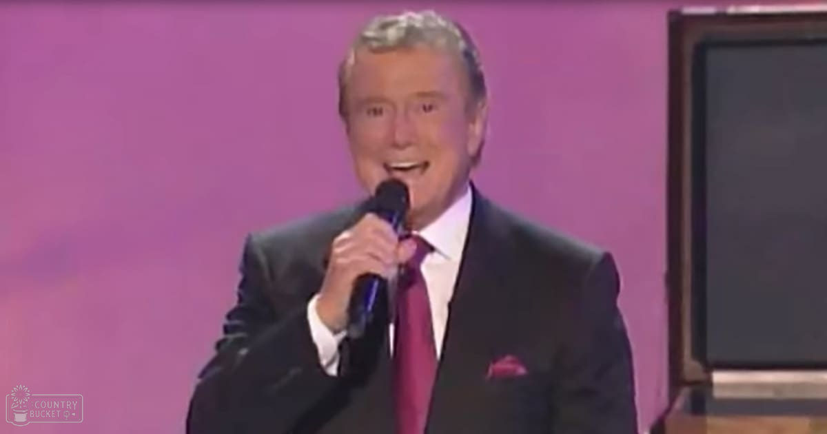 A middle-aged man wearing a dark suit, white shirt, and red tie stands in front of a solid pink backdrop, suggesting an indoor performance or event setting.