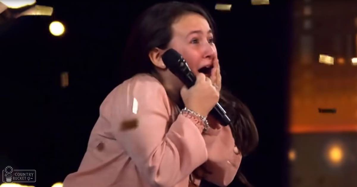A young adult female in a light pink blouse sings into a microphone on stage with blurred stage lights behind her, suggesting a performance setting.