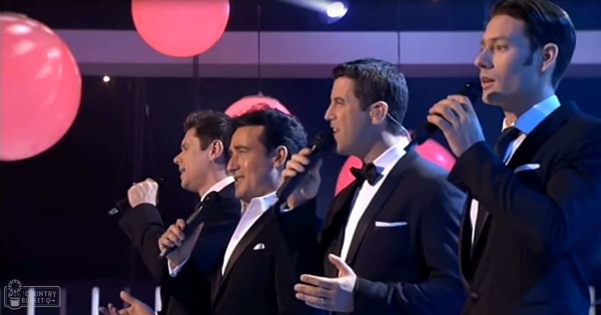 A group of male singers performing in a formal indoor setting with red spherical decorations hanging from above, illuminated by stage lighting.